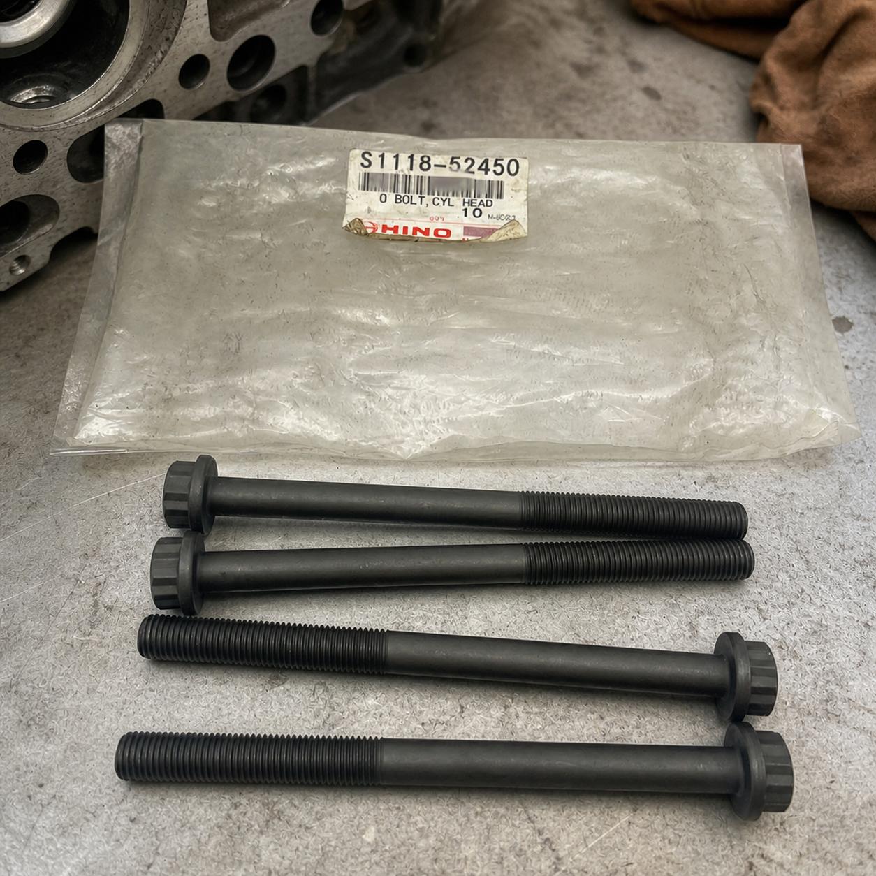 A front-view shot of four long, dark-grey engine bolts arranged horizontally, with a plastic bag containing a Hino-branded label resting behind them.